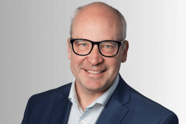 A professional head shot of a smiling man with glasses and a blue suit, resized to a 1 6: 9 aspect ratio.