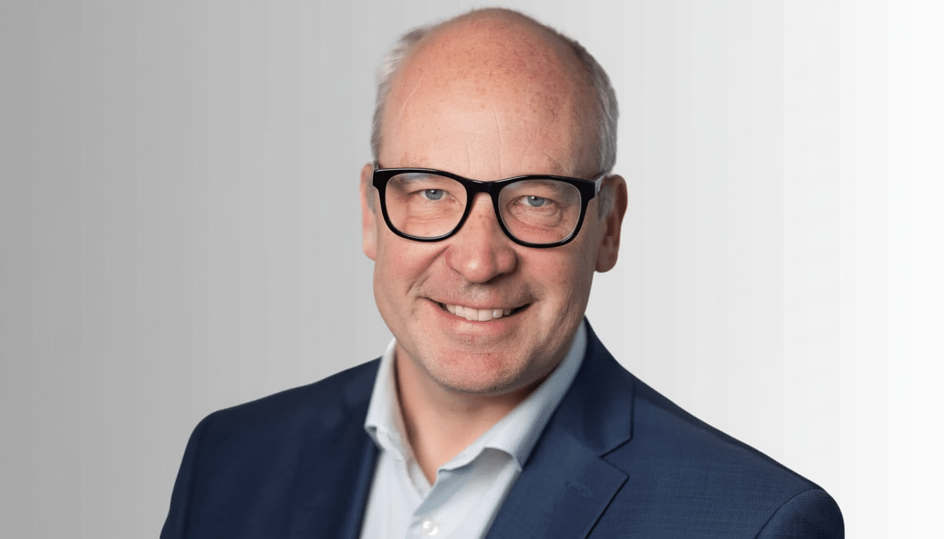 A professional head shot of a smiling man with glasses and a blue suit, resized to a 1 6: 9 aspect ratio.