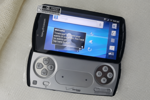 A top -down view of a Sony Ericsson Xperia Play phone with its gamepad controls slid out, resting on a textured light-colored surface . The screen displays a Welcome Times cape message and a Google search bar .