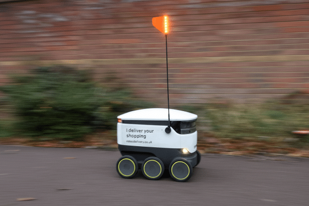 A white and black delivery robot with six wheels and an orange flag on top , moving on a paved path with a blurred brick wall and greenery in the background.