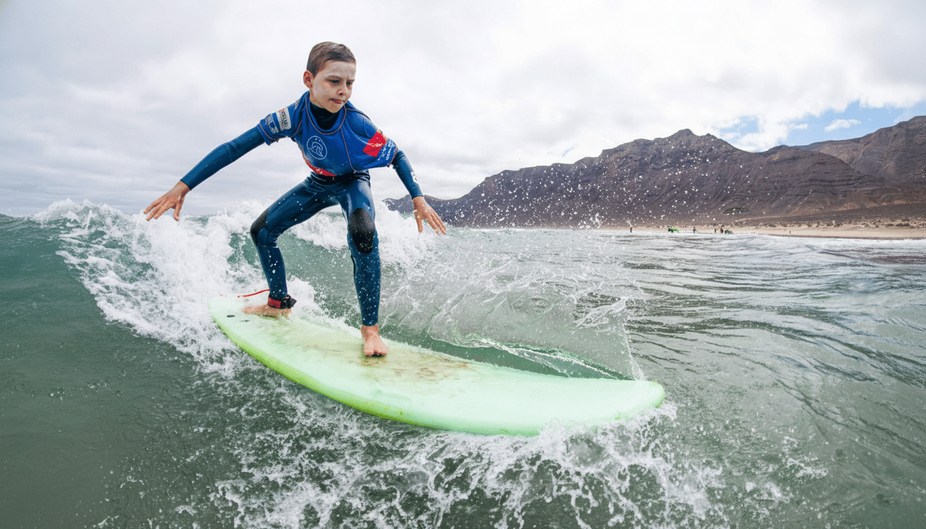 A young boy in a blue and red wetsuit sur fs on a green surfboard, riding a wave with mountains in the background .