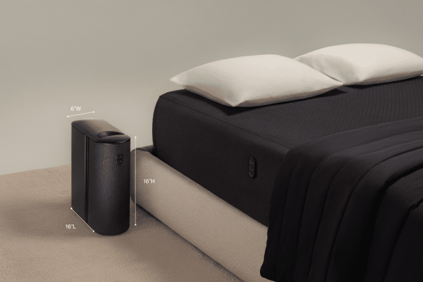 A dark gray rectangular air purifier with rounded edges and dimensions 6 W , 16 H, and 16 L, stands next to a bed with a dark gray mattress, white pillows , and a dark gray blanket. The background is a plain light gray wall and a light beige carpet .