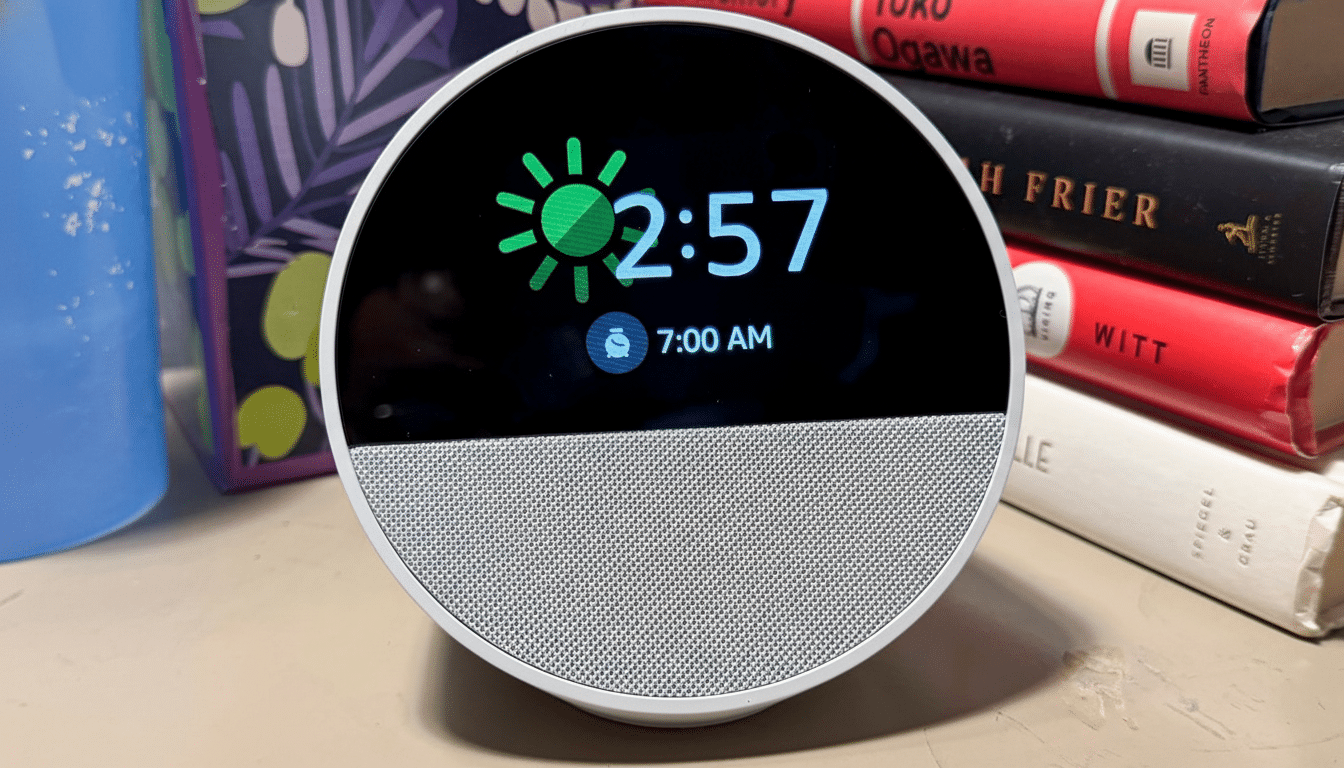 A white smart clock with a circular screen showing the time 2: 57, a sun icon, and a 7:0 0 AM alarm, set on a surface with books in the background.
