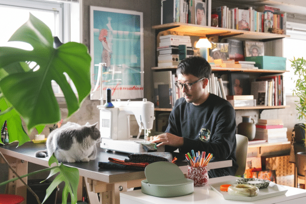 A man wearing glasses and a black sweatshirt is seated at a sewing machine , working on fabric , with a gray and white cat observing him from the desk. The room features bookshelves , plants , and natural light. Filename : man sewing withcat. png