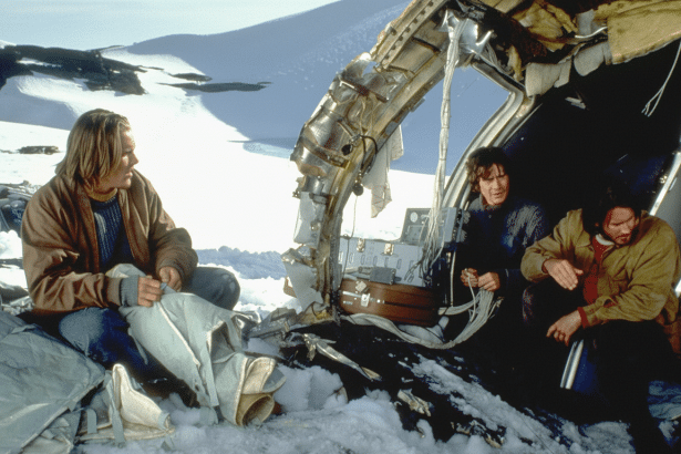 Three individuals , two men and one woman, sit amidst the wreckage of a plane in a snowy , mountainous landscape. One man on the left looks towards the other two, who are seated closer to the plane 's remains .