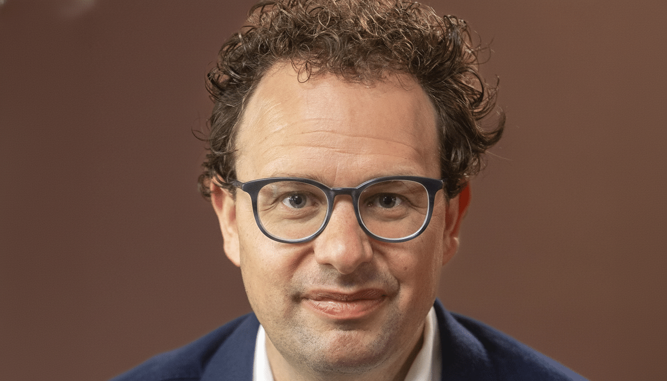 Head shot of a man with curly brown hair and glasses , smiling against a plain brown background .