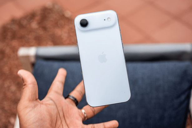 A hand holding a white iPhone with a custom camera bump, set against a blurred background of a textured surface and outdoor furniture, resized to a 16