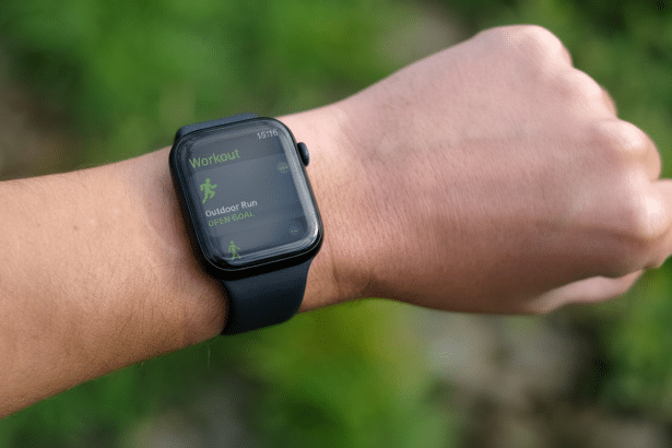 Close -up of a person's wrist wearing an Apple Watch displaying the Workout app , with Outdoor Run selected and Open Goal visible on the screen. The background is a natural , out -of-focus green.