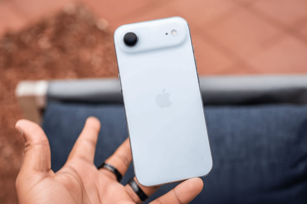 Hand holding a white iPhone with the Apple logo visible on the back, against a blurred outdoor background.