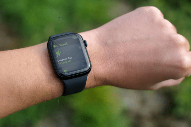 Close -up of a person's wrist wearing an Apple Watch , displaying the Workout app with Outdoor Run selected , set against a blurred green outdoor background. Filename : apple watchoutdoor runworkout .png