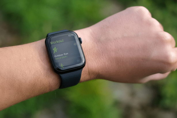 Close -up of a persons wrist wearing an Apple Watch, displaying the Workout app with Outdoor Run selected, set against a blurred green background.