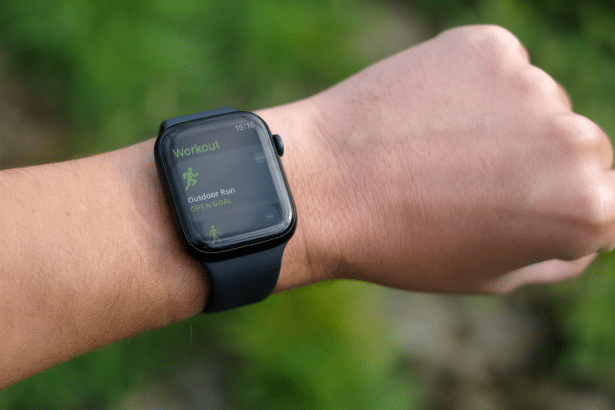 Close -up of a persons wrist wearing an Apple Watch displaying the Workout app, with Outdoor Run selected and OPEN GOAL visible against a blurred gree