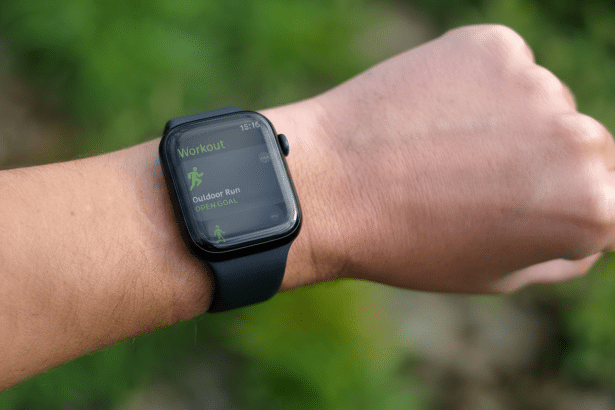 Close -up of a person's wrist wearing an Apple Watch , displaying the Workout app with Outdoor Run selected , set against a blurred green background .