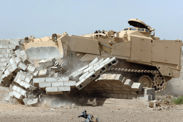 A desert camouflage armored personnel carrier is actively demolishing a concrete block wall, with debris and dust flying around the front of the vehicle.