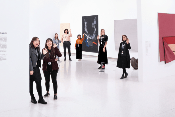 Several people stand in a brightly lit art gallery, with some waving towards the viewer. Various artworks are displayed on the white walls.