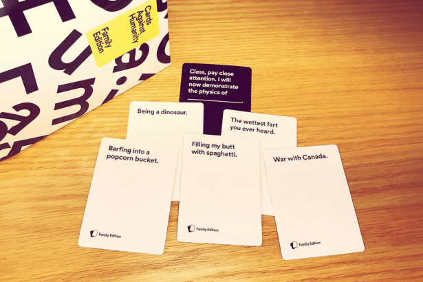 An overhead view of a wooden table with several Cards Against Humanity Family Edition cards laid out. One black card reads Class, pay close attention. I will now demonstrate the physics of and several white answer cards show humorous phrases. A partially visible Cards Against Humanity Family Edition box is in the upper left corner. Filename : cardsagainst humanity familyedition 16x 9. png