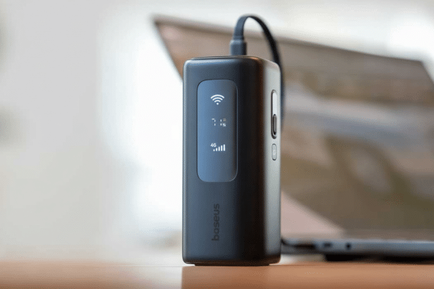 A black Baseus portable Wi -Fi hotspot device , displaying signal strength and battery life , sitting on a wooden desk with a laptop blurred in the background, resized to 1 6:9 aspect ratio.