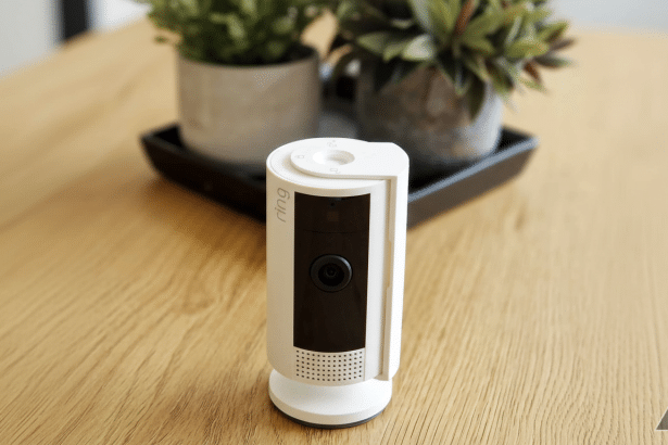A white Ring security camera standing on a wooden surface, with two potted plants in the blurred background.