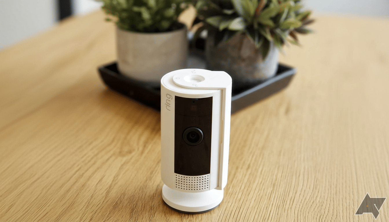 A white Ring security camera standing on a wooden surface, with two potted plants in the blurred background.
