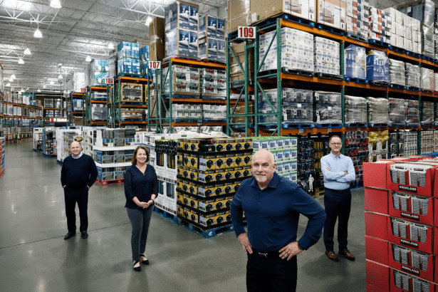 Four people standing in a warehouse with tall shelves stocked with various products.