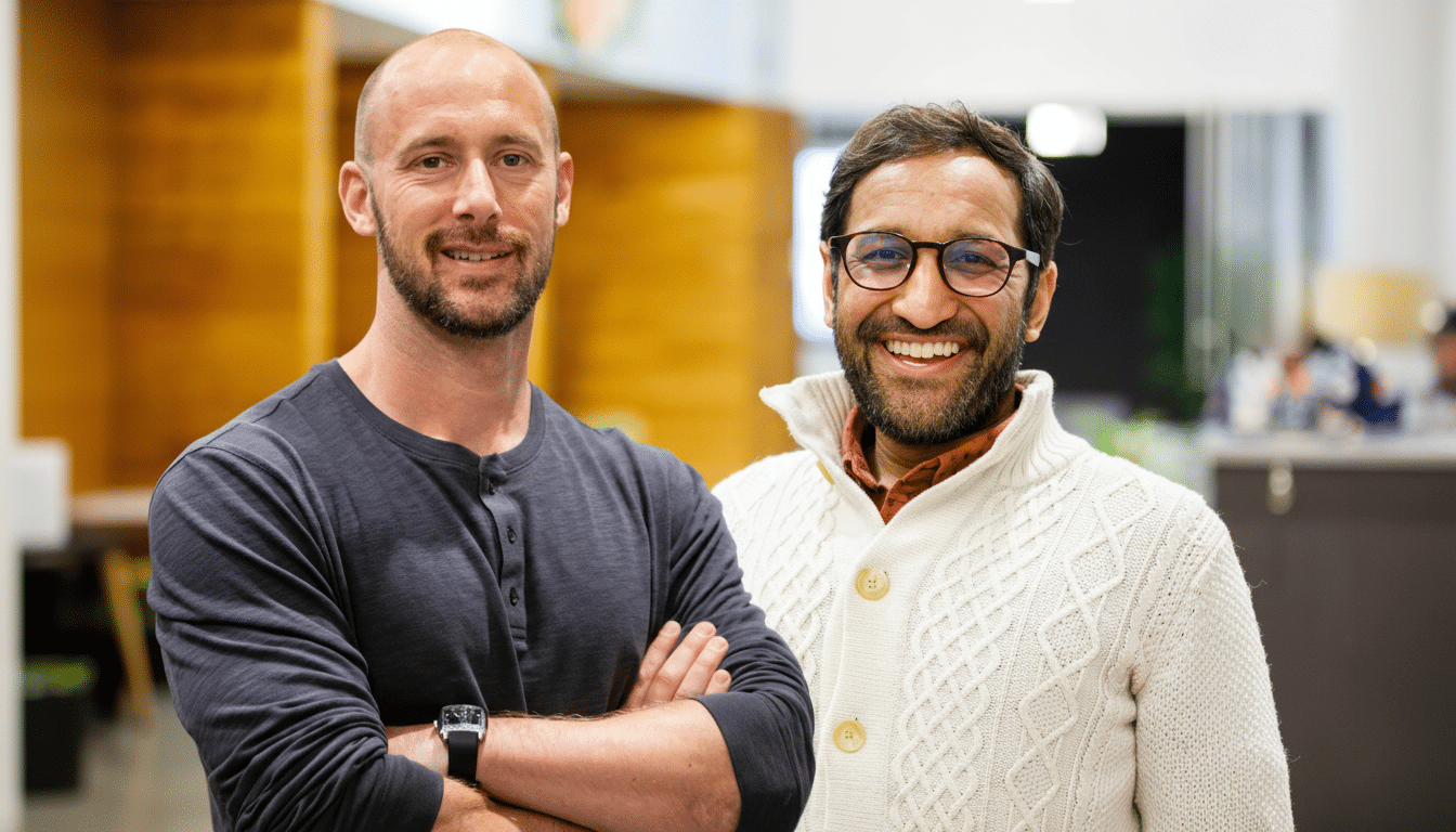 Two men, one with a beard and dark shirt, and another with glasses and a white cardigan, smiling in an office setting.