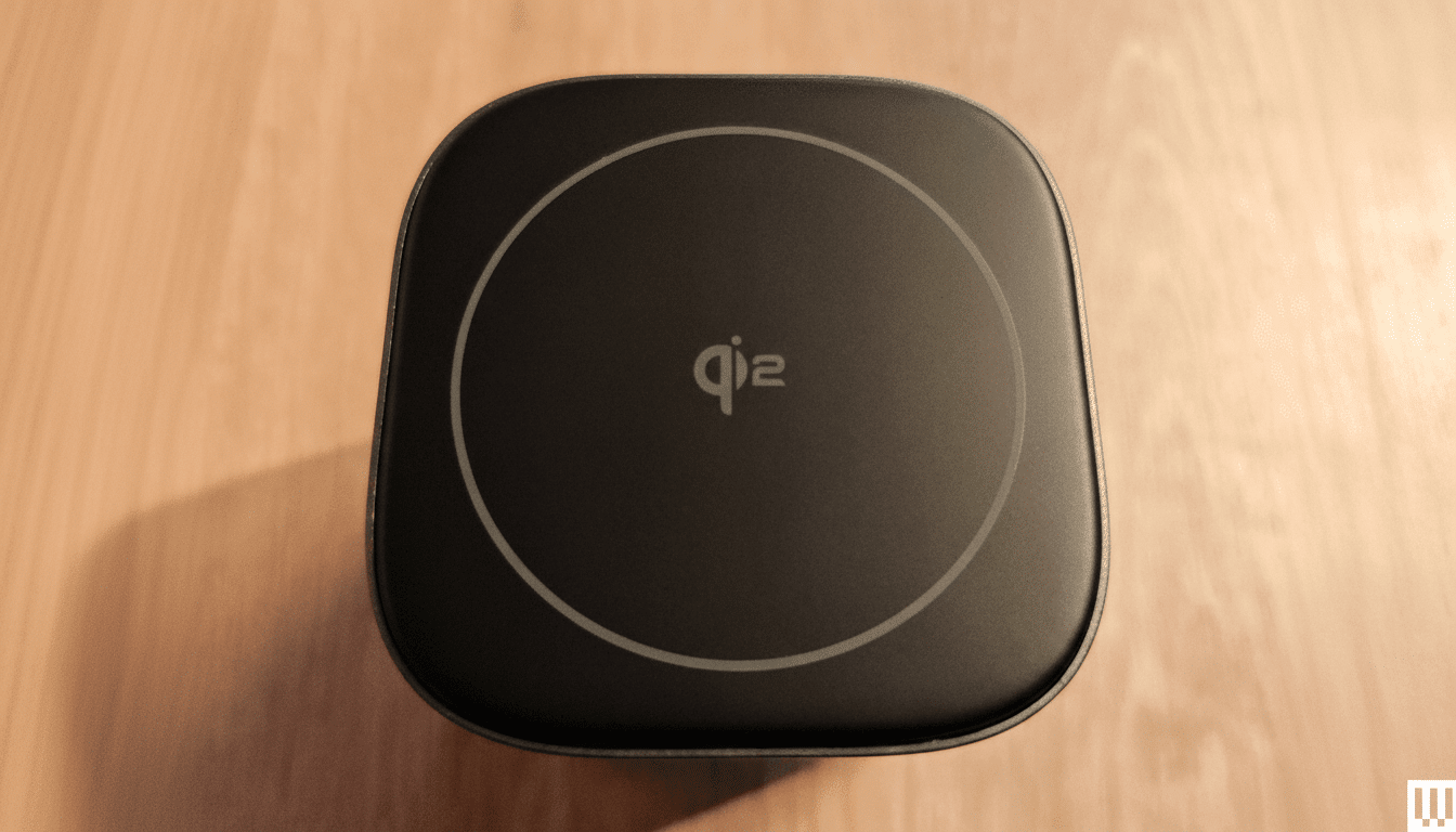 A top -down view of a black square wireless charger with the qi 2 logo and a white circular outline on its surface, set against a wooden background.