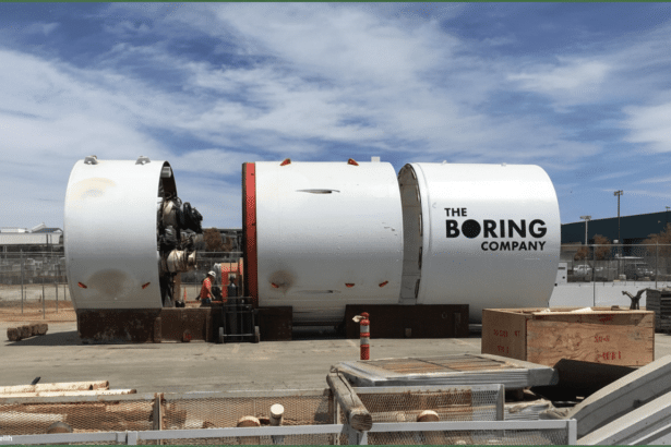 The Boring Company's tunneling machine, consisting of three large white cylindrical sections, sits outdoors under a clear sky , with a worker in the middle section. Filename : boringcompany tunneling machine.png
