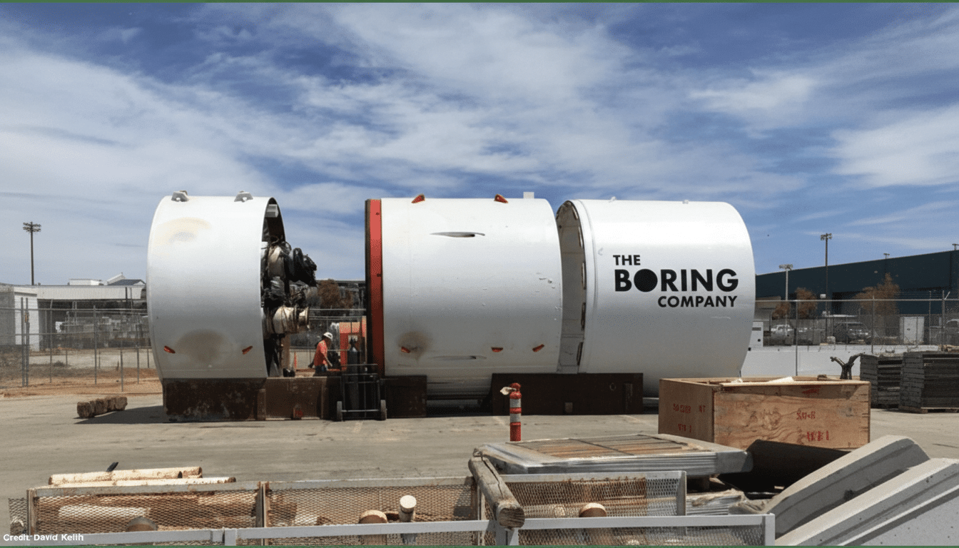 The Boring Company's tunneling machine, consisting of three large white cylindrical sections, sits outdoors under a clear sky , with a worker in the middle section. Filename : boringcompany tunneling machine.png