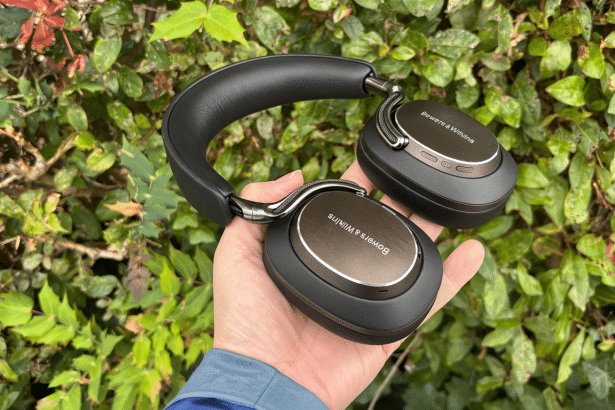 A hand holds a pair of black Bowers & Wilkins over -ear headphones against a lush green leafy background. Filename : bowers wilkinsheadphones hand resized .png
