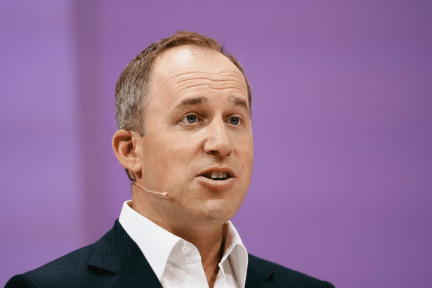 A man in a dark suit and white shirt, wearing a lavalier microphone, speaks against a soft purple background. His gaze is directed slightly upward and
