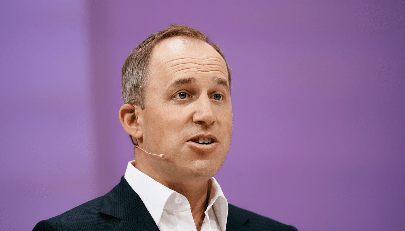 A man in a dark suit and white shirt, wearing a lavalier microphone, speaks against a soft purple background. His gaze is directed slightly upward and
