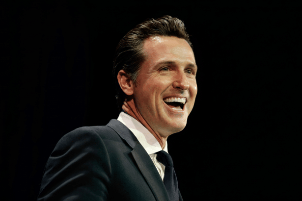 A man with dark hair in a suit and tie , smiling broadly against a dark background, resized to a 16: 9 aspect ratio.