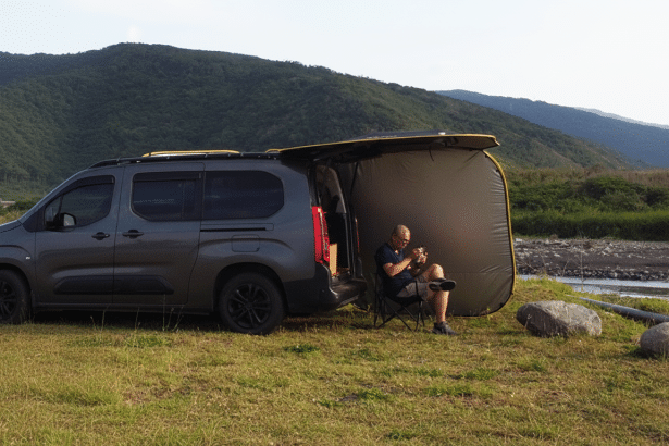 A man sitting in a camping chair next to a dark grey van with an attached awning , set up in a grassy field beside a river with mountains in the background , resized to 16 :9 aspect ratio .