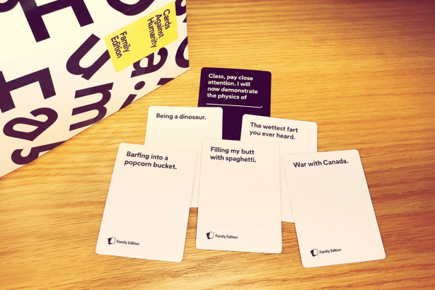 An overhead shot of a Cards Against Humanity Family Edition game box and several white and one black game cards spread out on a wooden table. Filename : cardsagainst humanity familyedition game. png