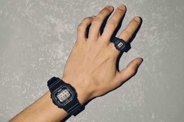 A hand wearing a black G -Shock wristwatch and a matching G -Shock ring, against a textured gray background, resized to 1 6:9 aspect ratio. Filename : hand with gshock watches 1 69. png