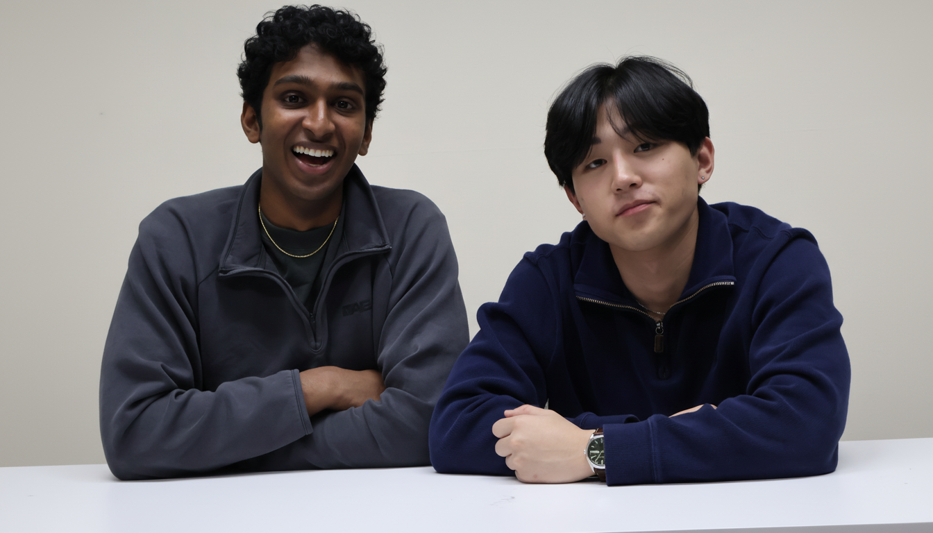 Two young men, one with dark curly hair and a wide smile, and the other with straight dark hair and a neutral expression, are seated at a white table against a light background.