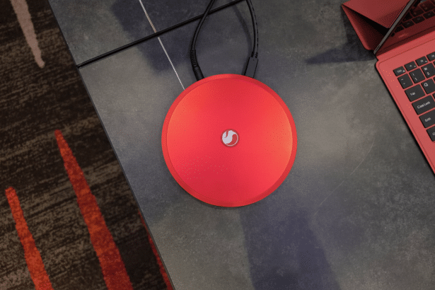 A red circular device with a white logo, connected to two black cables, sitting on a dark grey surface next to a red laptop keyboard.