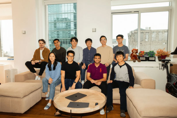 A group of eleven young adults, appearing to be a team or startup, smiling and posing in a modern, well -lit living room setting with laptops on a cof
