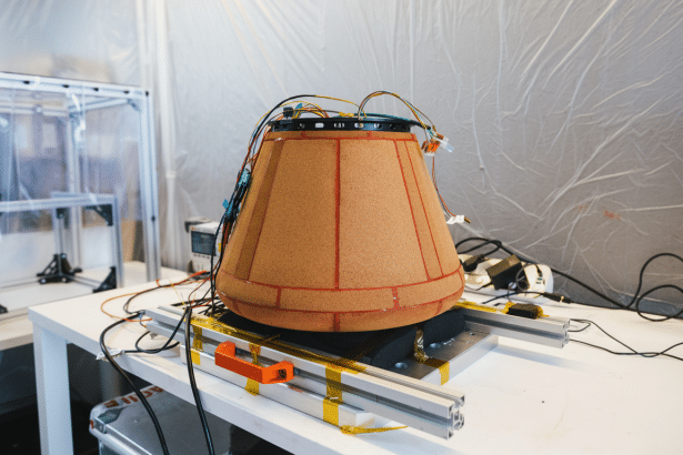 A conical object covered in cork- like material and wired with electronics , situated on a metallic rail system on a white table. Filename : conical electronic device ontable .png