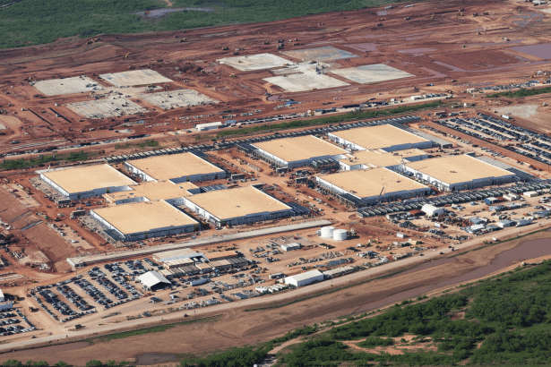 Aerial view of a large construction site with multiple square buildings and vast areas of cleared red earth, surrounded by some green vegetation.