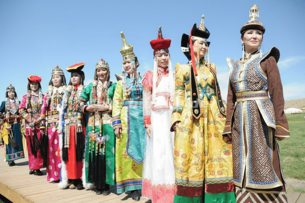 Several individuals dressed in traditional, colorful Mongolian attire stand in a line on a wooden platform under a clear blue sky. Filename : mongolian att ireline. png