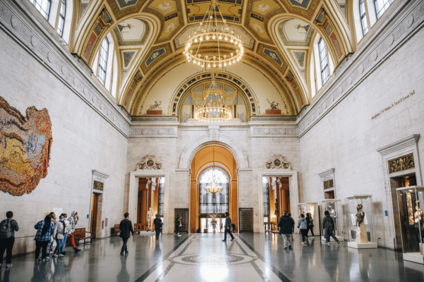 The Grand Hall of the Philadelphia Museum of Art, featuring a high arched ceiling with ornate gold and pale yellow decorations , large chandeliers, and marble walls. Several people are walking through the hall, admiring the exhibits. Filename : phil adelphiamuseum grandhall. png