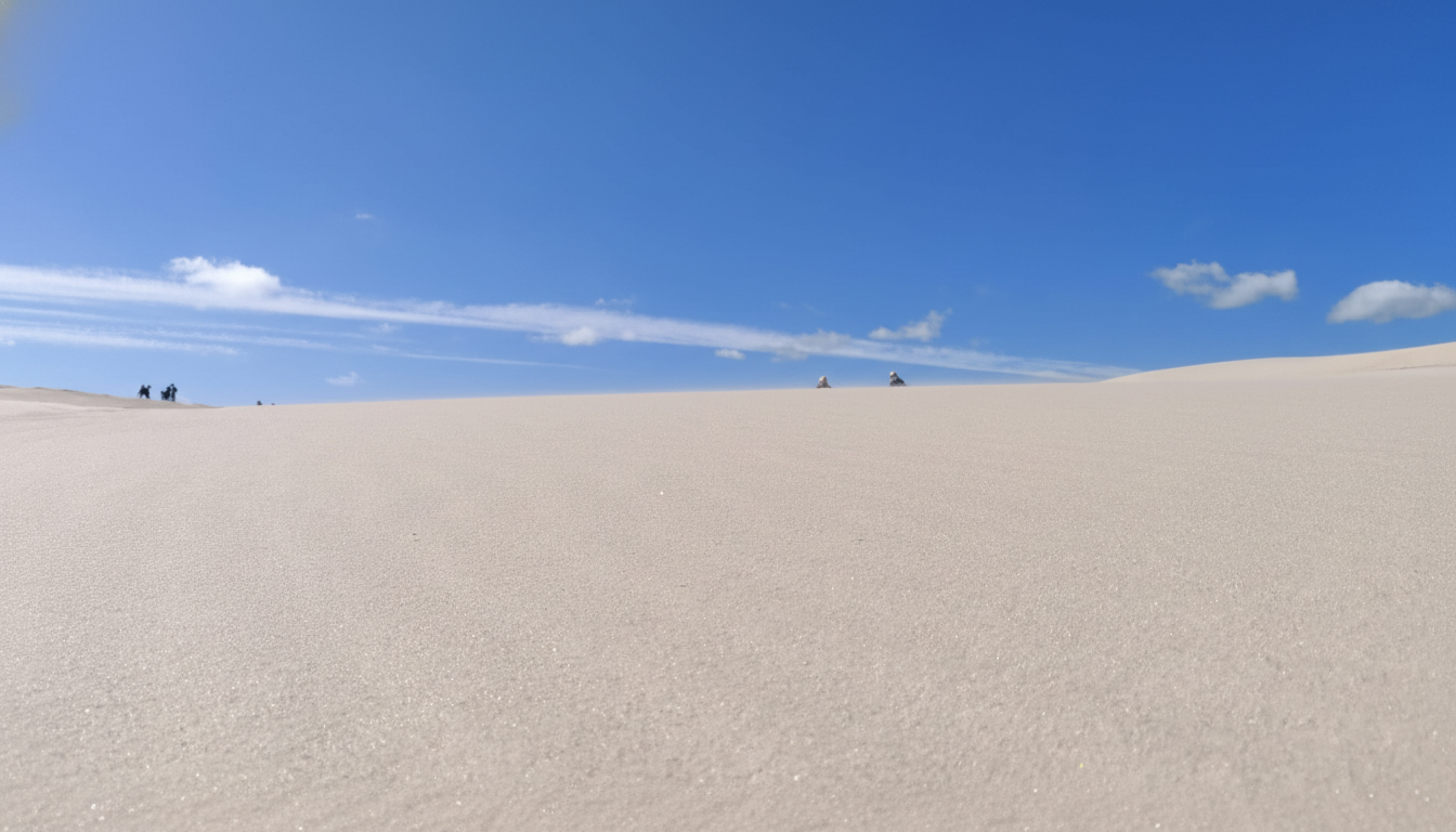 A wide shot of a vast sand dune under a bright blue sky with scattered clouds, with a few small figures visible in the distance on the left horizon .