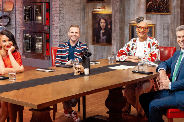 Four smiling judges , two men and two women, seated at a long wooden table with a black runner, with ovens and framed art in the background.