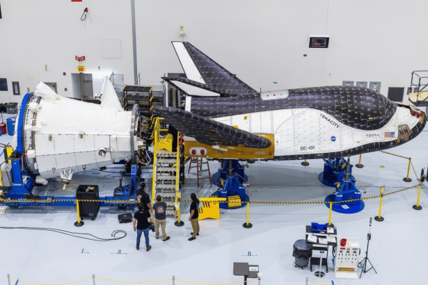 An aerial shot of the Dream Ch aser spacecraft Tenacity during assembly , showing its white and black body , with engineers working around it in a brightly lit hangar.