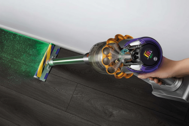 A hand holding a Dyson vacuum cleaner, with its green laser detecting dust on a dark wooden floor.