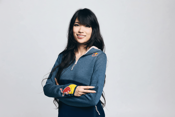 A young woman with long dark hair, wearing a blue and yellow Red Bull branded hoodie, smiling at the camera with her arms crossed.