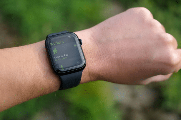 A close -up shot of a person's wrist wearing an Apple Watch , displaying the Workout app with Outdoor Run and Open Goal selected . The background is a natural, out -of-focus green. Filename : applewatch workoutapp . png