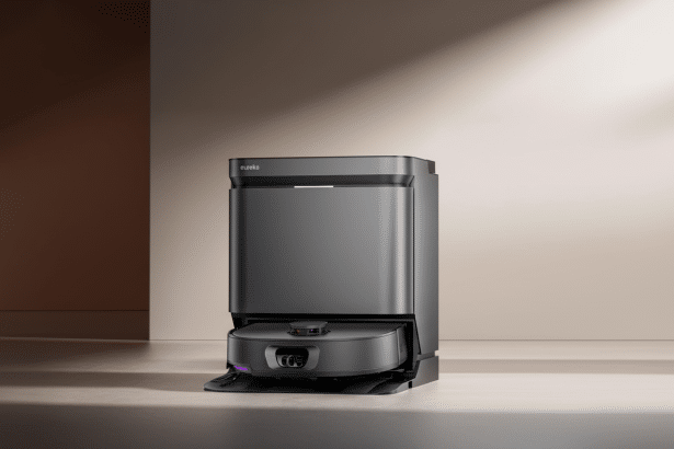A dark gray Eureka robot vacuum cleaner docked in its charging station , set against a two -tone background with warm neutral colors and subtle lighting .