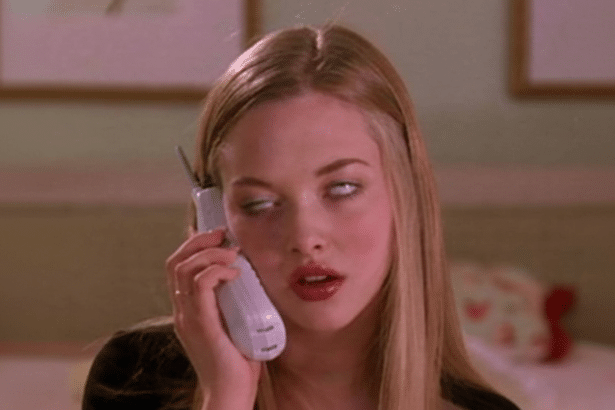 A young woman with blonde hair holds a white flip phone to her ear , rolling her eyes upwards with an exasper ated expression. The background is a soft pink room.